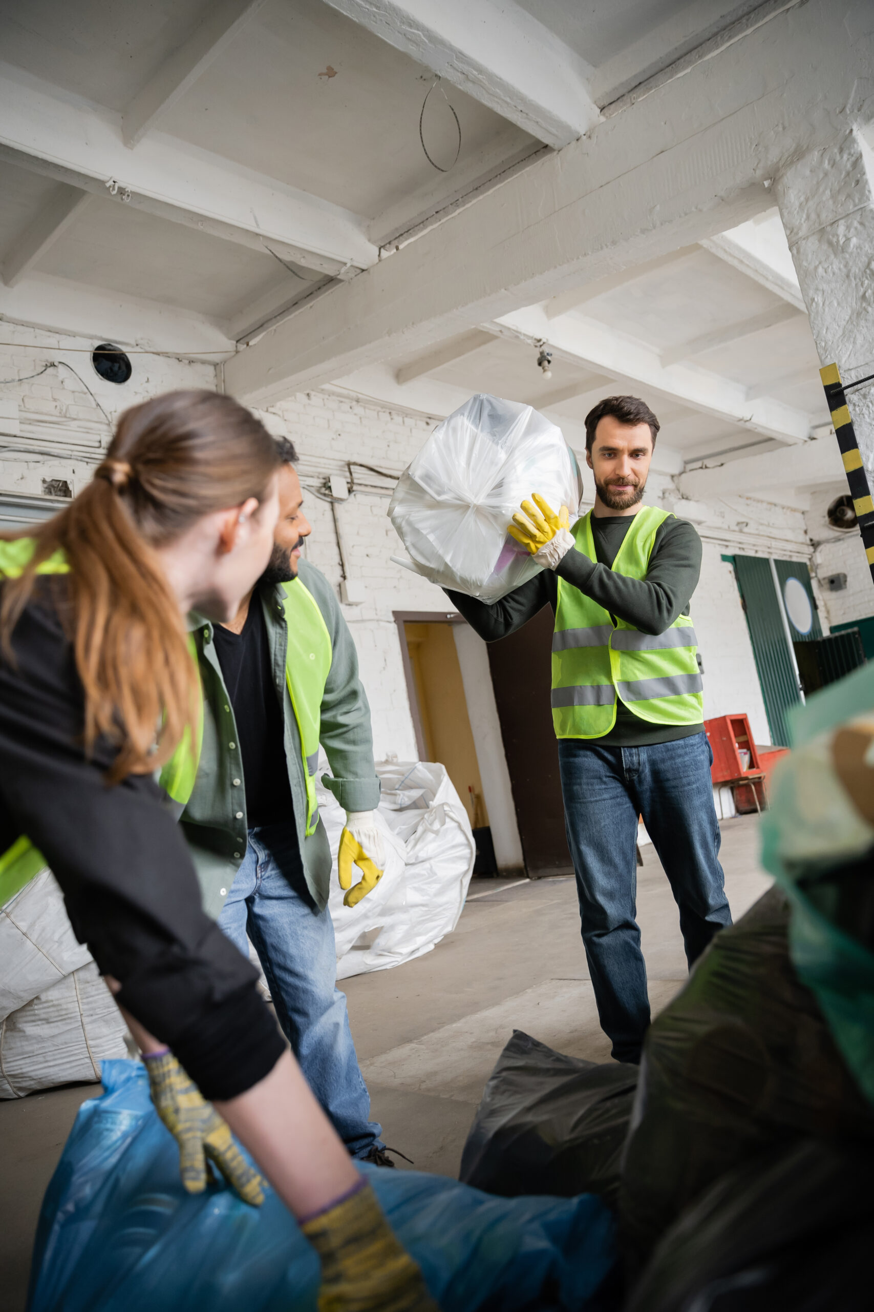Worker in protective vest and glove holding plastic bag with trash near blurred interracial colleagues working in waste disposal station, garbage sorting and recycling concept